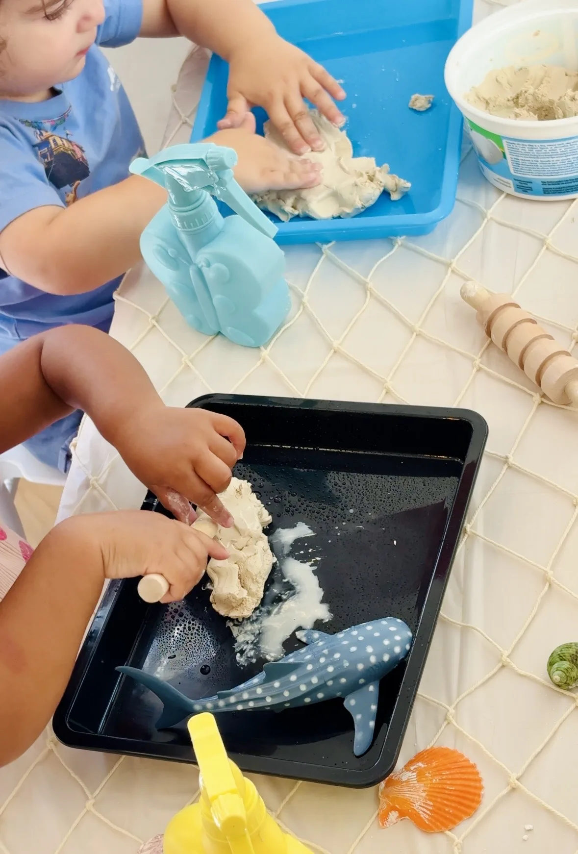 Toddlers exploring an ocean-themed sensory tray with kinetic sand, shells, and a whale shark